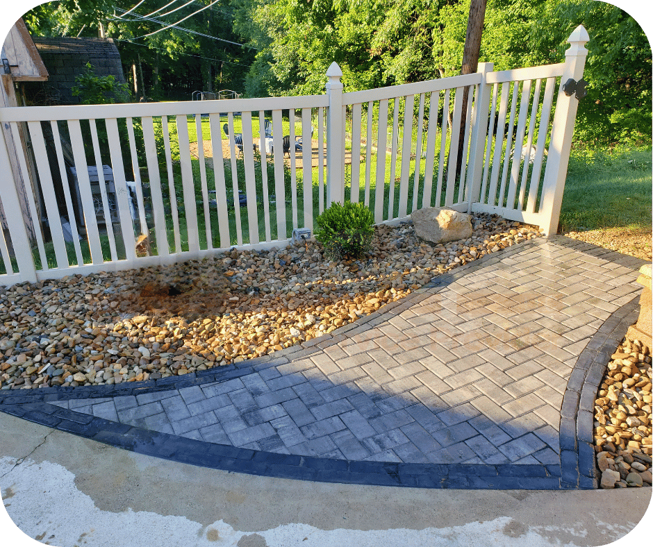 Paved walkway bordered by decorative rocks and a small shrub, enclosed by a white picket fence in a backyard garden.