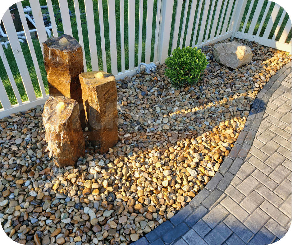 Small backyard water feature with three stone pillars surrounded by decorative rocks, next to a white fence and paver walkway.