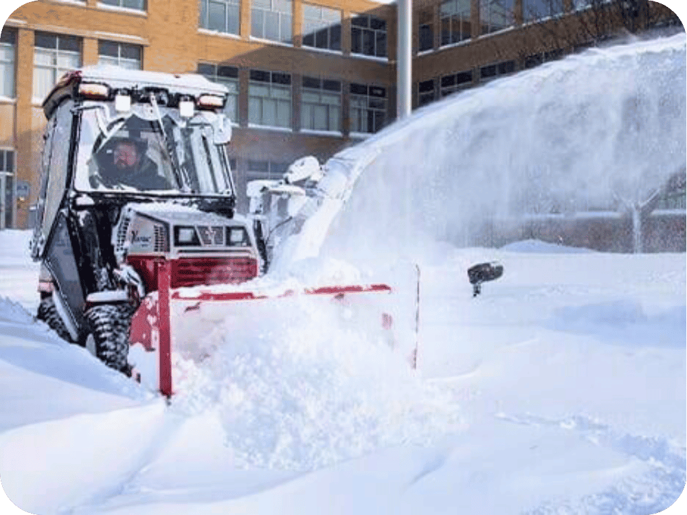 Worker operating a snow blower to clear snow from a parking lot in front of a building.