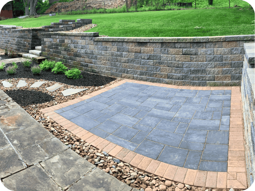 Stone patio area with brick accents, surrounded by retaining walls, decorative rocks, and a landscaped garden featuring small shrubs and stepping stones.