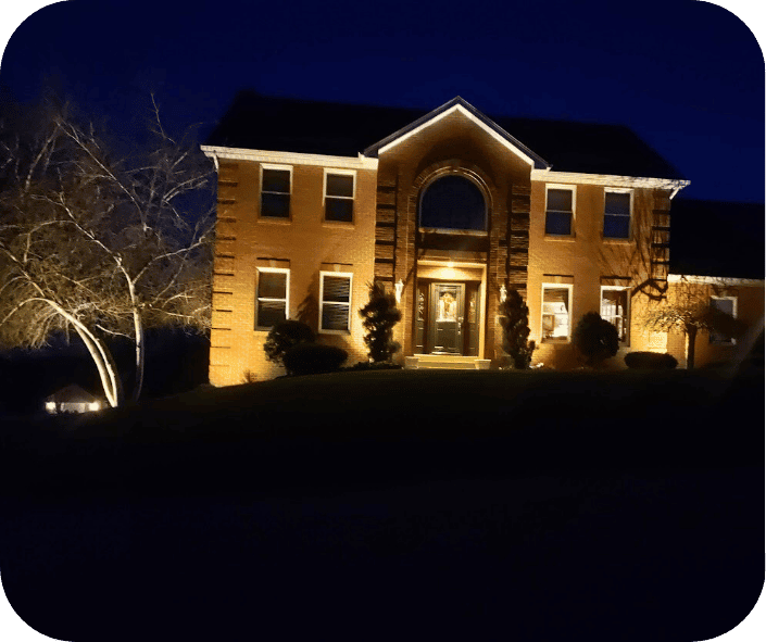 Brick house illuminated by outdoor lighting at night, with trees and shrubs accentuated by the warm glow.