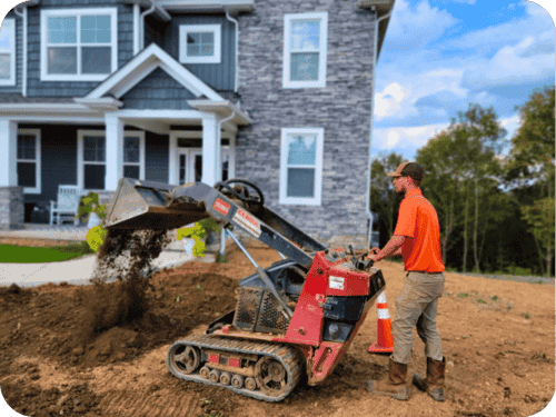 A Uniformed TruScape employee in an orange polo shirt operating a walk behind skid steer spilling dirt. 