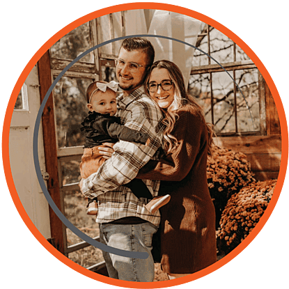 Happy family of three, parents holding their baby daughter, standing in front of a rustic background.