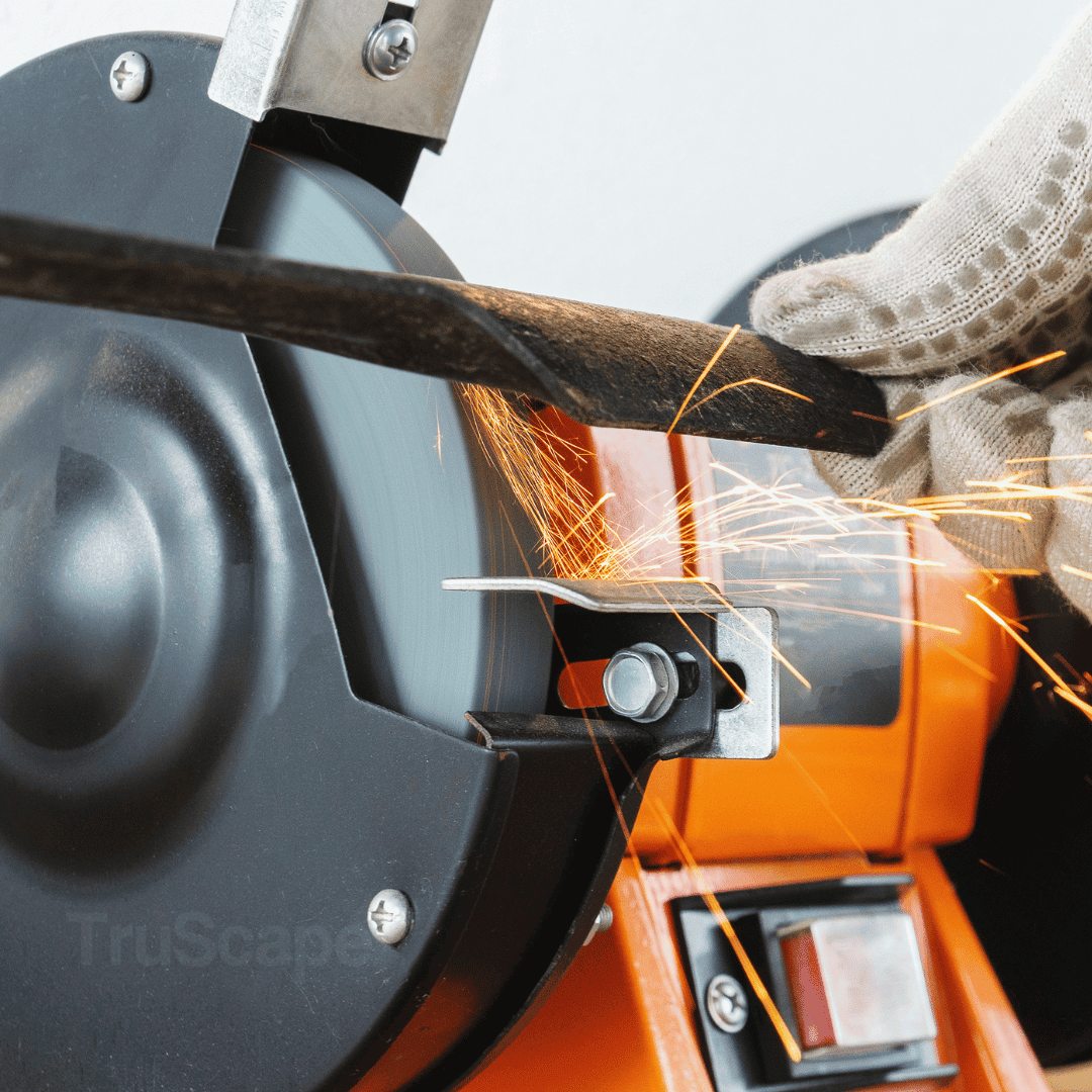 Close-up of a lawnmower blade being sharpened on a bench grinder, with sparks flying as a gloved hand holds the blade steady.