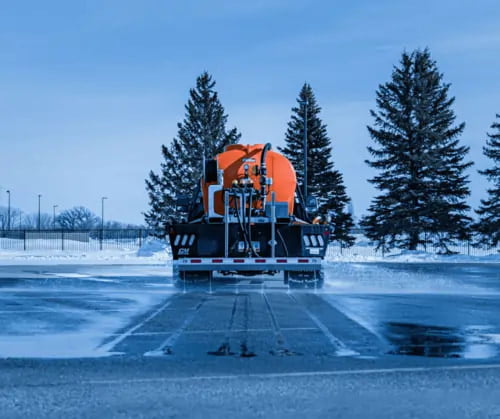 A truck with an orange de-icing tank sprays brine solution onto a snow-covered parking lot, with evergreen trees in the background.