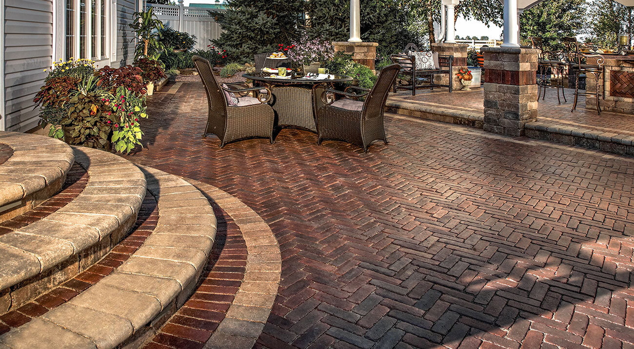 Elegant backyard patio with red brick herringbone pavers, curved stone steps, outdoor dining set, and surrounding planters with lush greenery.