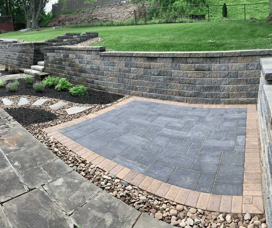 Landscaped backyard with stone retaining walls, a paved patio area, stepping stones, and small shrubs surrounded by mulch and decorative rocks.