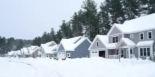 A suburban neighborhood covered in snow, with houses lined up along a quiet street and snowbanks piled up from plowing.