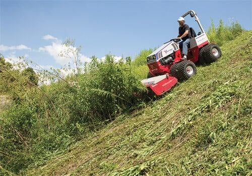 Man operating a red Ventrac mower on a steep grassy hillside, clearing overgrown vegetation.