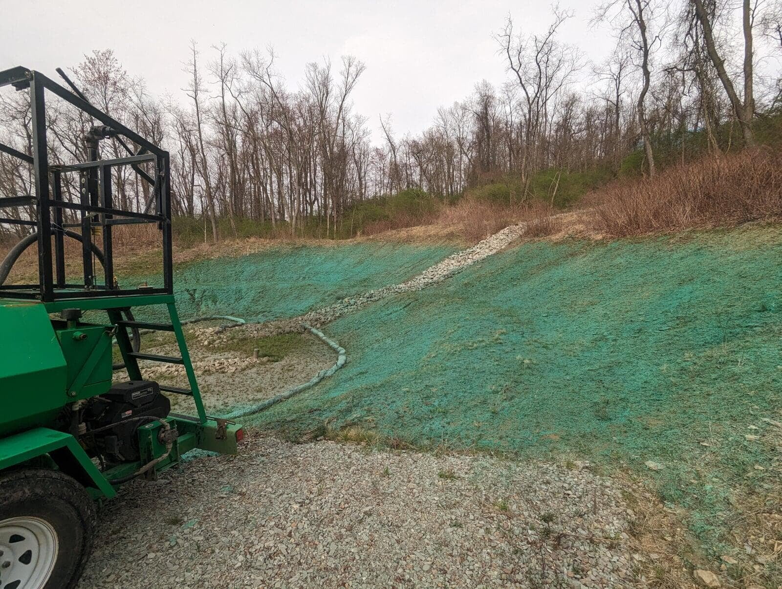 A barren hillside freshly covered with green hydroseeding material to promote grass growth. A hydroseeding machine is partially visible in the foreground, and a gravel pathway cuts through the slope. The background shows leafless trees and brush along the horizon.