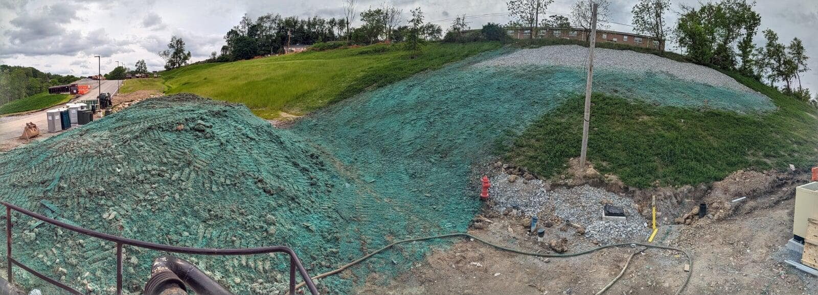 A wide view of a construction area where a large sloped hill is covered with hydroseeding material. In the distance, a grass field and trees are visible. A fire hydrant and gravel are situated near the bottom of the hill, with utility lines and construction equipment also present.