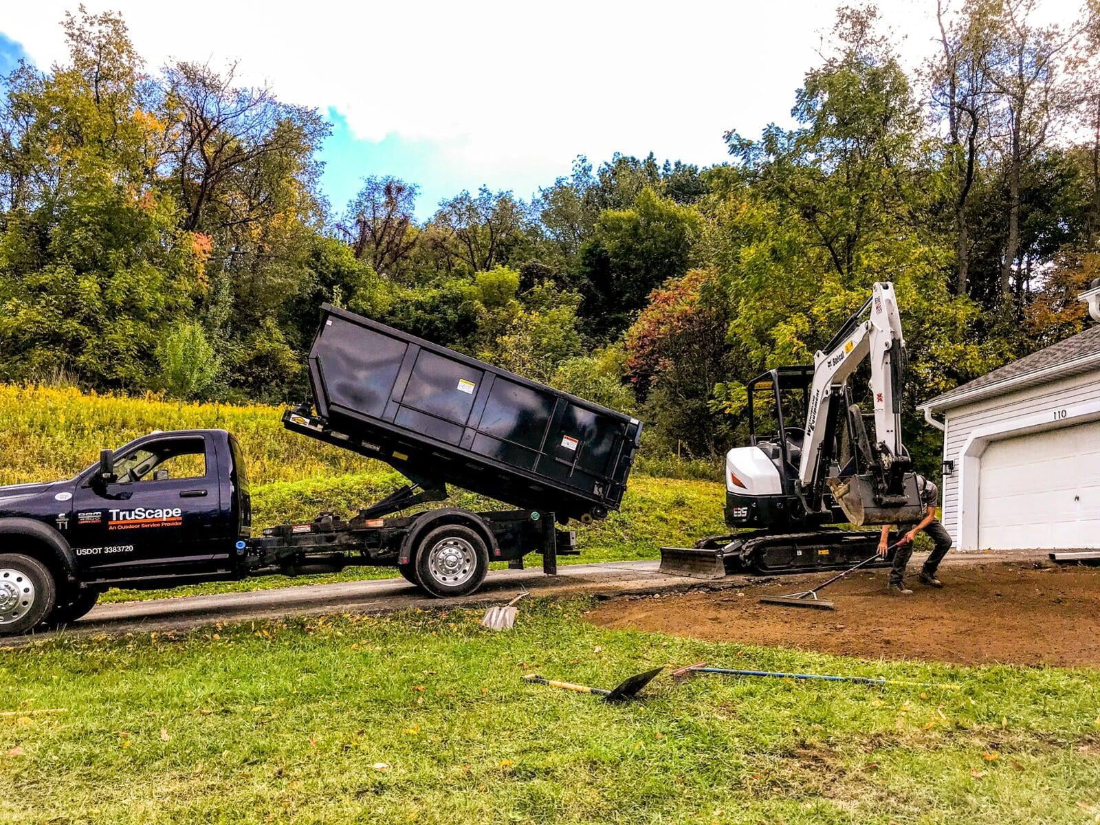 An excavator removes topsoil from a nearby dump truck while an employee spreads with a rake. 