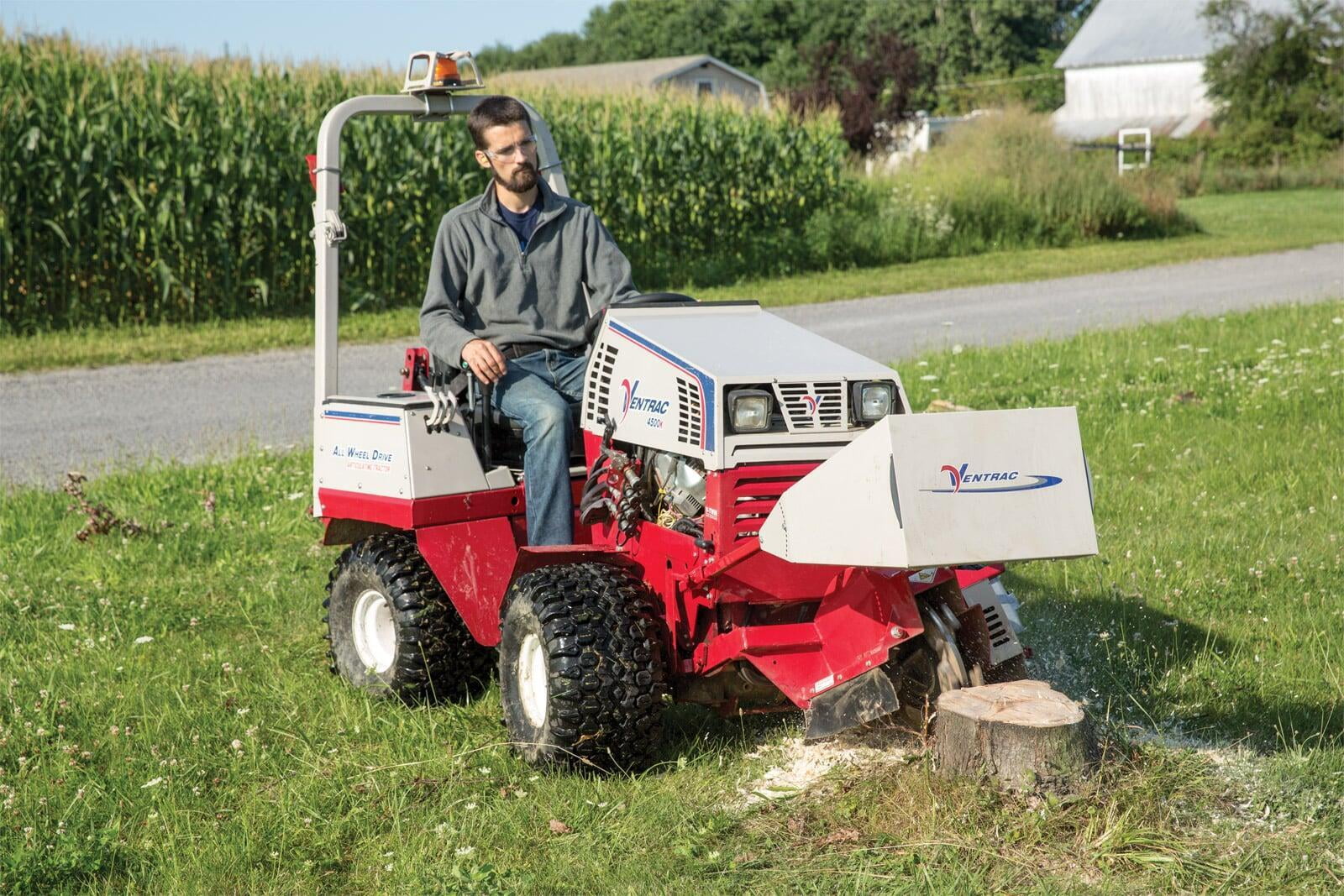 Man using a Ventrac 4500 tractor with a stump grinder attachment to remove a tree stump in a grassy area near a cornfield.