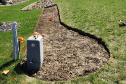 Freshly prepared garden bed with soil cleared for landscaping, bordered by grass, next to a utility box with marker flags.