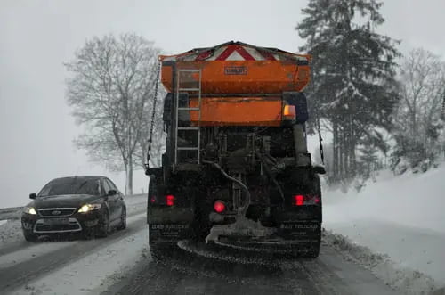 A salt truck spreads de-icing material on a snow-covered road during a snowstorm, with a car passing by on the left.