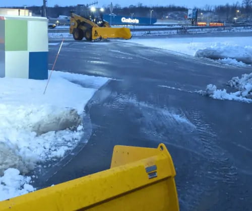 A snowplow clears snow from a parking lot near a retail store in the early evening, with piles of snow along the edges of the pavement.