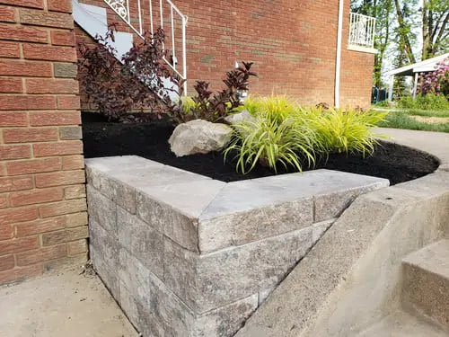 Stone retaining wall with a small garden bed featuring ornamental grass and shrubs beside a brick house.