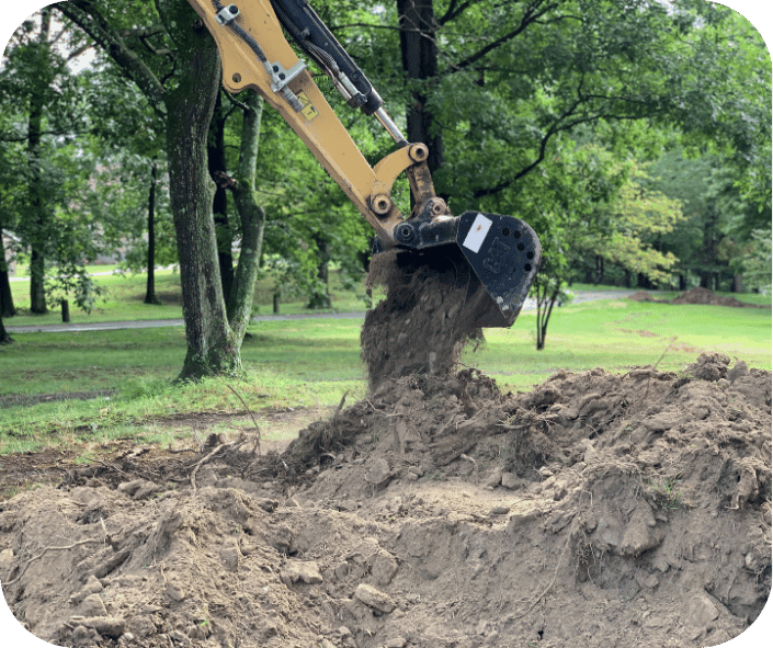 Excavator digging soil in a grassy area with trees in the background, preparing land for construction or landscaping.