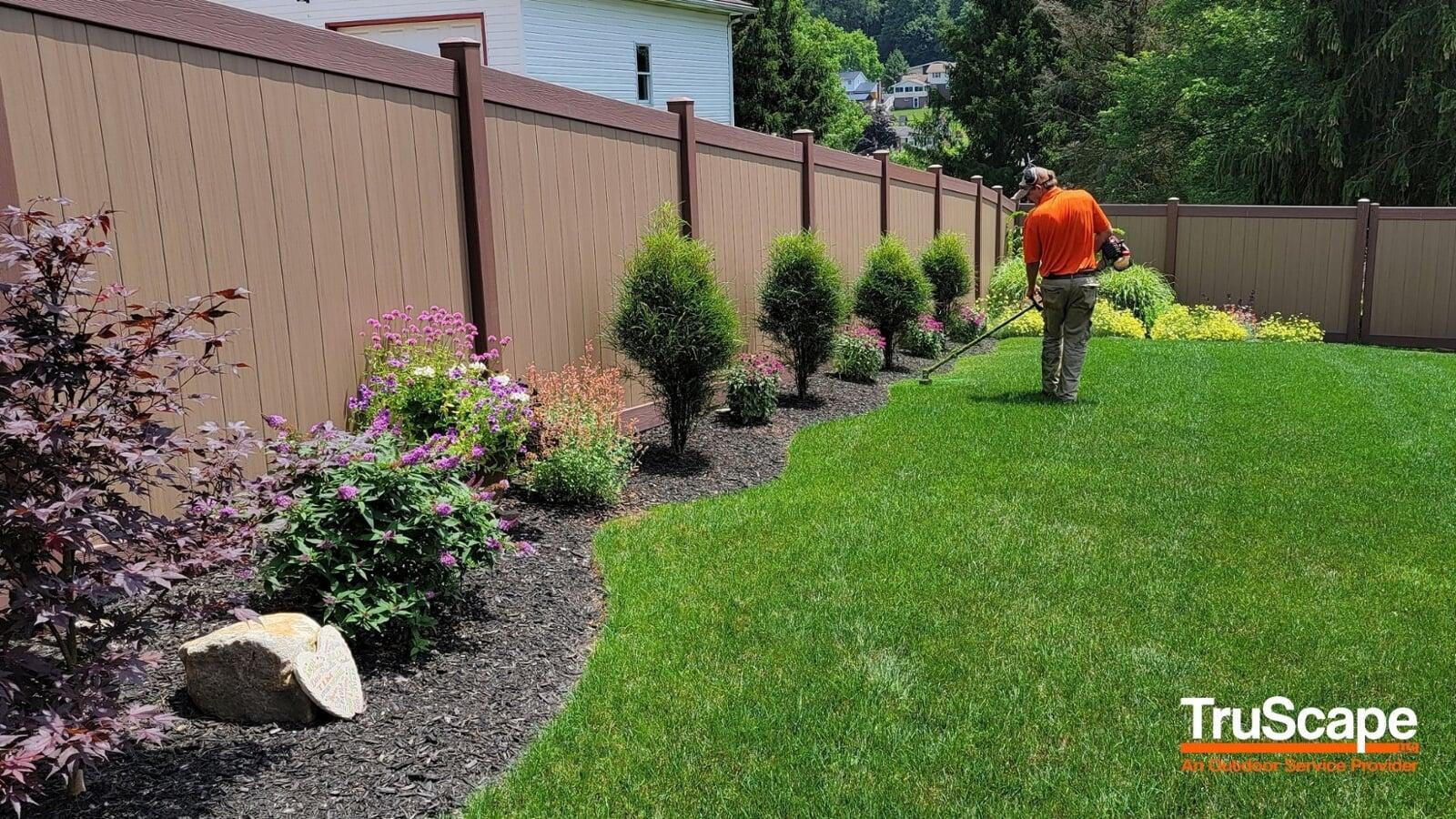 Landscaper trimming grass along a flower bed with blooming plants and small shrubs, bordered by a brown privacy fence in a well-maintained backyard.