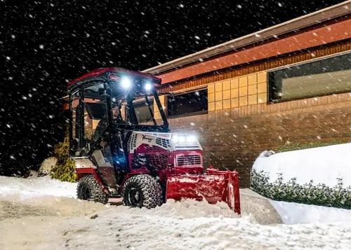 A snow plow operator clearing a sidewalk at night during heavy snowfall, with bright headlights illuminating the scene.