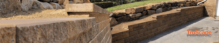 Versa-Lok retaining wall made of tan concrete blocks, supporting a sloped garden with neatly stacked stone. TruScape logo visible in the corner.