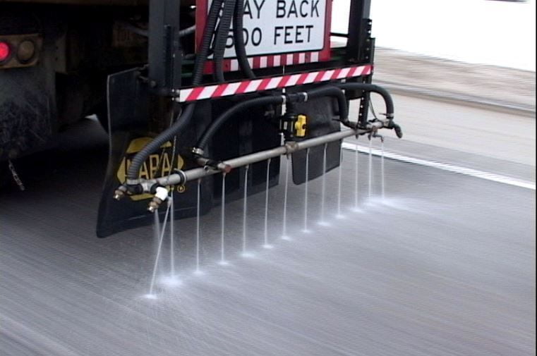 A de-icing truck spraying a liquid solution onto a road surface, with a "Stay Back 100 Feet" sign on the rear.