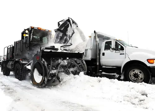 A snow blower truck loading snow into a dump truck during a snow removal operation on a winter day.